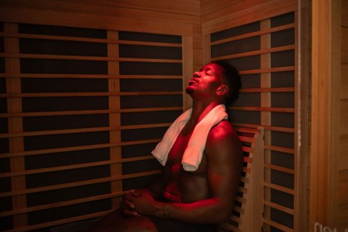 A man sits in a sauna, eyes closed, with a towel around his neck, illuminated by soft red light.