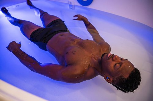 A person floating in a bath with blue lighting, eyes closed, appearing relaxed and serene.