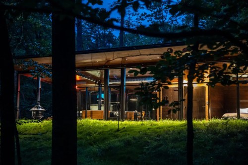 A modern cabin illuminated at night, surrounded by trees and greenery, showcasing large glass windows and a cozy atmosphere.
