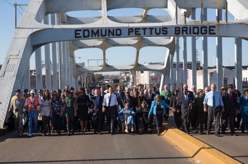 A diverse group of people marches across the Edmund Pettus Bridge, commemorating a historic civil rights event.