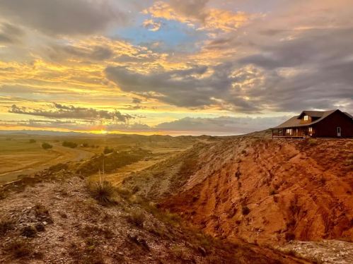 A rustic house on a hillside at sunset, with vibrant clouds and a vast landscape in the background.