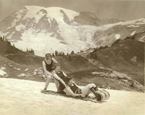 Three women in vintage swimsuits sledding down a snowy slope with a mountain backdrop.