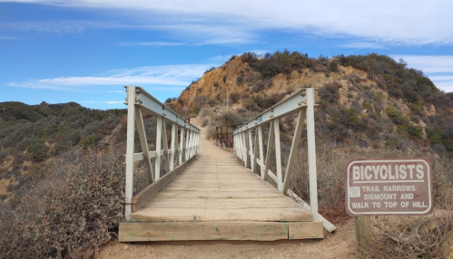 A wooden bridge leads to a hill, with a sign warning cyclists to dismount and walk to the top.