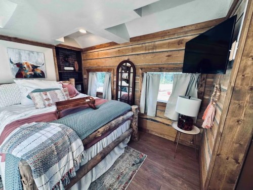 Cozy bedroom with wooden walls, a large bed, decorative pillows, a TV, and a rustic lamp. Natural light from windows.