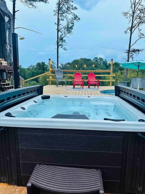 A hot tub on a deck surrounded by trees, with two red chairs and an umbrella in the background under a cloudy sky.