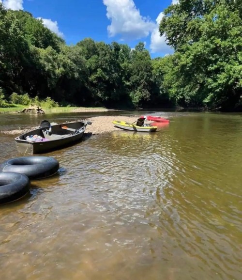 A serene river scene with boats and kayaks anchored near a sandy shore, surrounded by lush green trees under a blue sky.