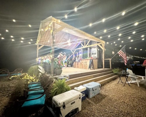 A cozy outdoor venue with string lights, a bar, and people enjoying a gathering by the water at night.