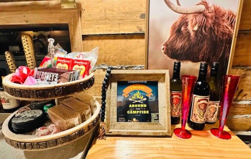 A rustic display featuring s'mores ingredients, a campfire sign, and decorative bottles on a wooden table.
