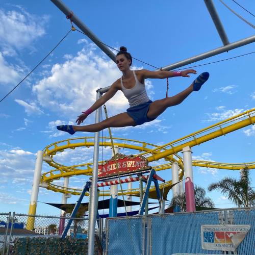 A performer executes a split on a trapeze against a blue sky, with amusement park rides in the background.