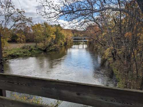 A serene river flows through a landscape of autumn trees under a cloudy sky, viewed from a wooden bridge.