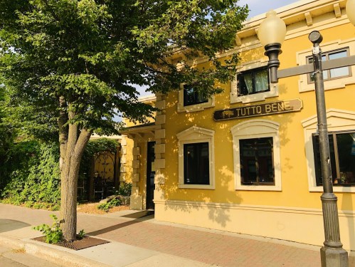 A yellow building with the sign "TUTTO BEN," surrounded by greenery and a tree, on a sunny day.