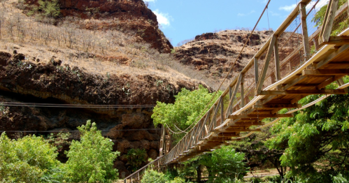 A wooden suspension bridge spans a lush green area, surrounded by rocky hills under a clear blue sky.