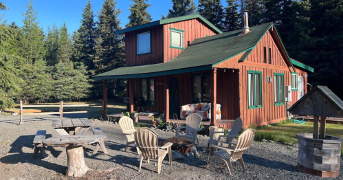A cozy red cabin surrounded by trees, featuring a porch and outdoor seating area with wooden chairs and a fire pit.