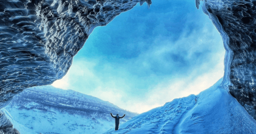 A person stands in an icy cave, arms raised, with a bright blue sky visible through the opening above.