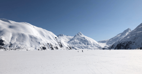 A vast snowy landscape with towering mountains under a clear blue sky, featuring two skiers in the distance.