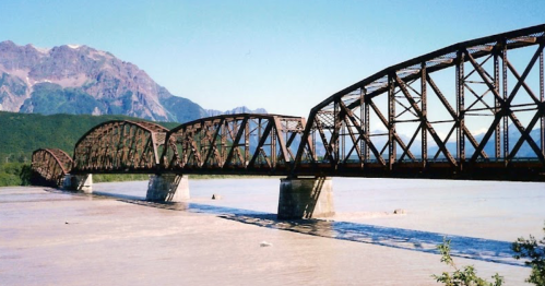 A rusted metal bridge spans a wide river, with mountains in the background under a clear blue sky.