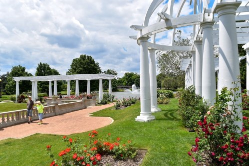 Two people stroll through a lush garden with white columns, colorful flowers, and a pond under a cloudy sky.
