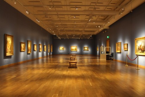 A spacious art gallery with wooden floors, blue walls, and framed paintings displayed evenly along the walls.
