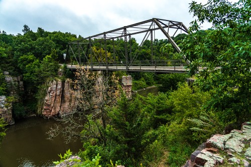 A metal bridge spans a river surrounded by lush greenery and rocky cliffs under a cloudy sky.