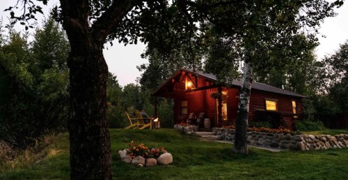 A cozy wooden cabin illuminated at dusk, surrounded by trees and a flower garden, with two chairs outside.