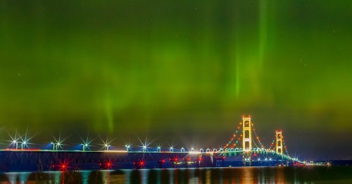 A vibrant aurora borealis illuminates the sky above a brightly lit bridge, reflecting on the water below.