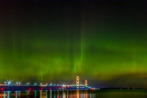 A vibrant aurora borealis illuminates the night sky above a bridge, reflecting in the calm water below.