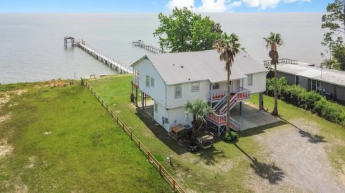 A waterfront house with a pier, surrounded by greenery and palm trees, overlooking a calm body of water.