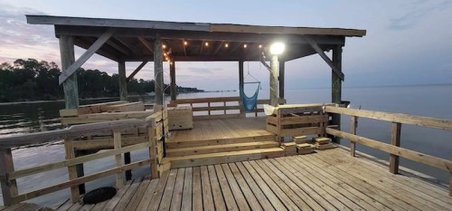 A wooden dock with a covered seating area, overlooking calm water at dusk, with soft lights illuminating the space.