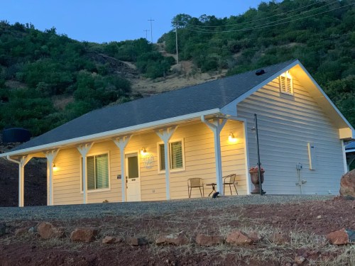 A cozy yellow house with a porch, set against a hillside at dusk, illuminated by warm outdoor lights.