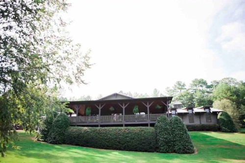 A large wooden house with a porch, surrounded by lush greenery and neatly trimmed hedges under a bright sky.