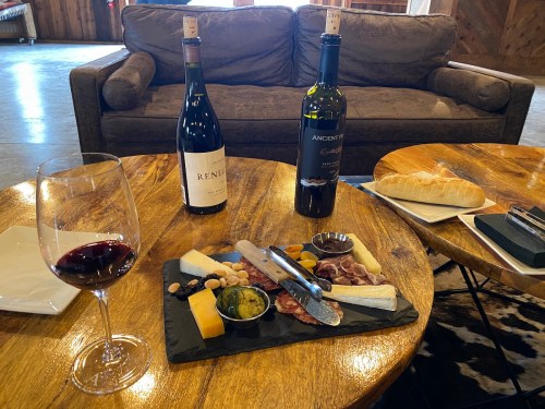 A wooden table with a charcuterie board, two bottles of wine, a glass of red wine, and a loaf of bread.