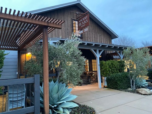 A rustic wine café with a wooden exterior, surrounded by greenery and a cloudy sky.