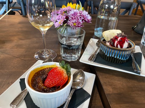 A table setting with a glass of white wine, a flower vase, and two desserts: crème brûlée with a strawberry and a sundae.