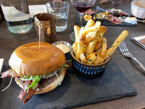 A gourmet burger with bacon and greens, served with a side of crispy fries in a blue cup on a slate plate.