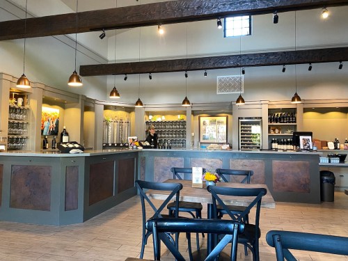 A modern tasting room with wooden beams, a bar, glassware, and seating, featuring warm lighting and a welcoming atmosphere.