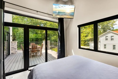 A modern bedroom with a TV, large windows, and a view of a deck with outdoor seating surrounded by trees.