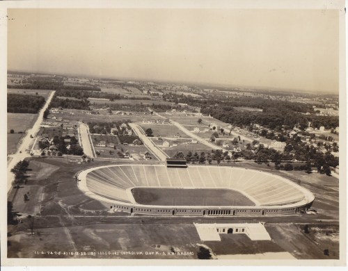 Aerial view of a large stadium surrounded by fields and residential areas, showcasing its circular structure and surrounding landscape.