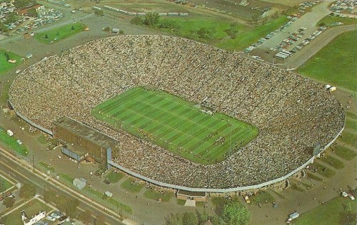 Aerial view of a packed stadium filled with fans, with a green field in the center and parking lots surrounding it.
