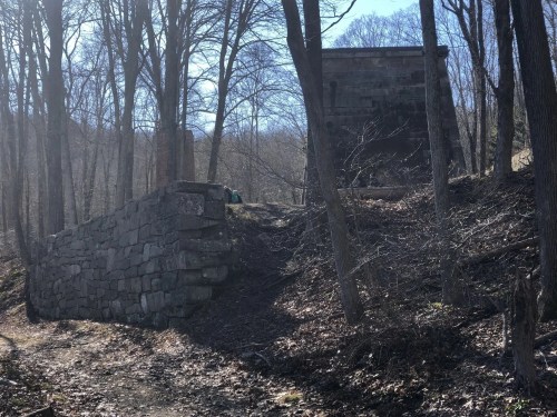 A stone wall and a partially collapsed structure surrounded by bare trees on a sunny day.