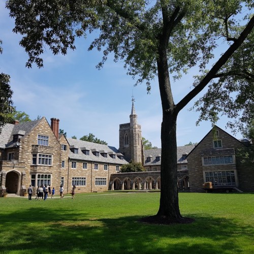 A stone building with a tower surrounded by green grass and trees, with people walking in the background on a sunny day.