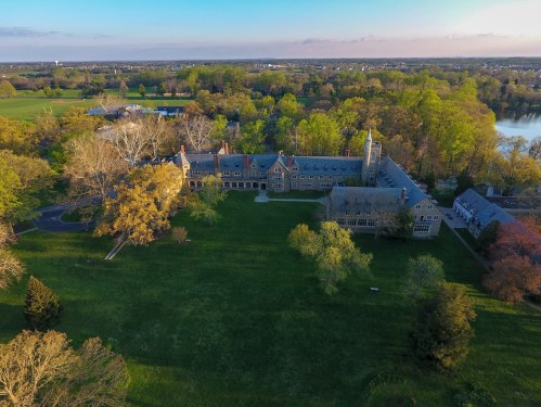 Aerial view of a large, historic building surrounded by lush greenery and trees, with a lake in the background.