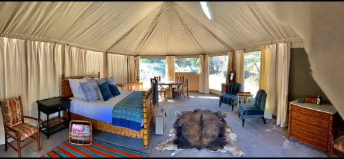 Cozy tent interior with a bed, dining area, and rustic decor, featuring a fur rug and large windows for natural light.