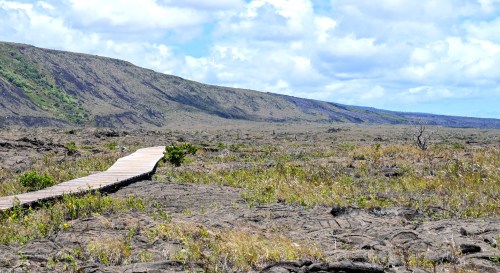 A wooden boardwalk stretches across a barren volcanic landscape under a blue sky with scattered clouds.