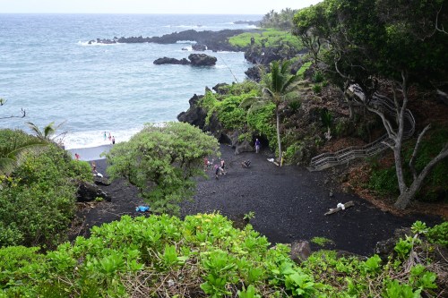A scenic view of a black sand beach surrounded by lush greenery and rocky coastline under a cloudy sky.