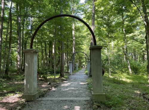 A serene forest path lined with stone pillars and arches, surrounded by lush green trees.