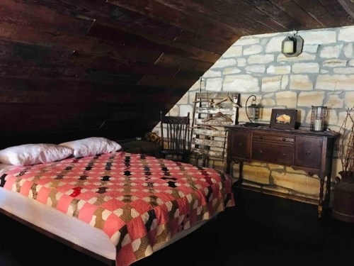 Cozy attic bedroom with a quilted bed, wooden furniture, and stone walls, featuring rustic decor and warm lighting.