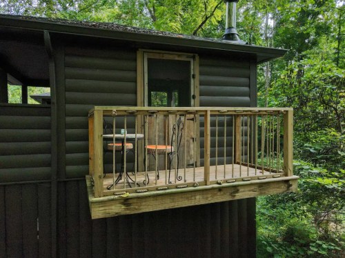 A small wooden balcony with two chairs overlooks a lush green forest, attached to a dark cabin.