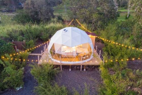 A geodesic dome house surrounded by greenery, with string lights illuminating the deck at dusk.