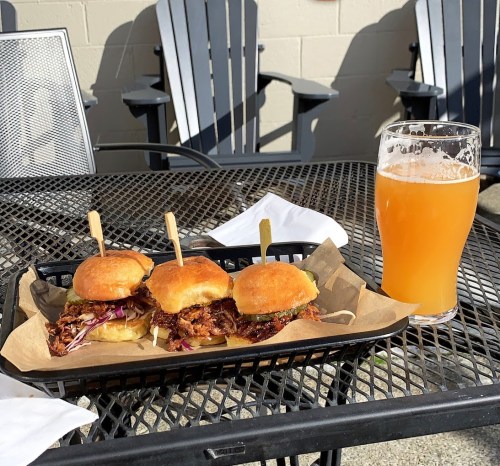 Three pulled pork sliders on a tray with a pint of orange beer on a black table outdoors.