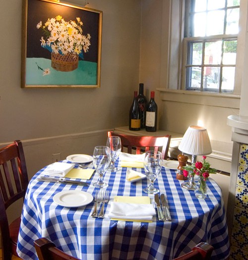 A cozy dining table with a blue checkered tablecloth, set with glassware and cutlery, near a flower painting and wine bottles.